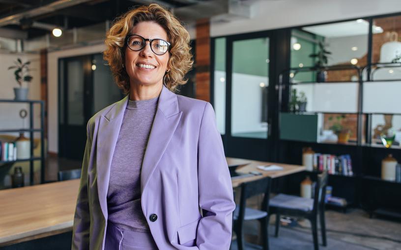 A person stands confidently in a business suit and smiles at you behind the camera in a meeting room