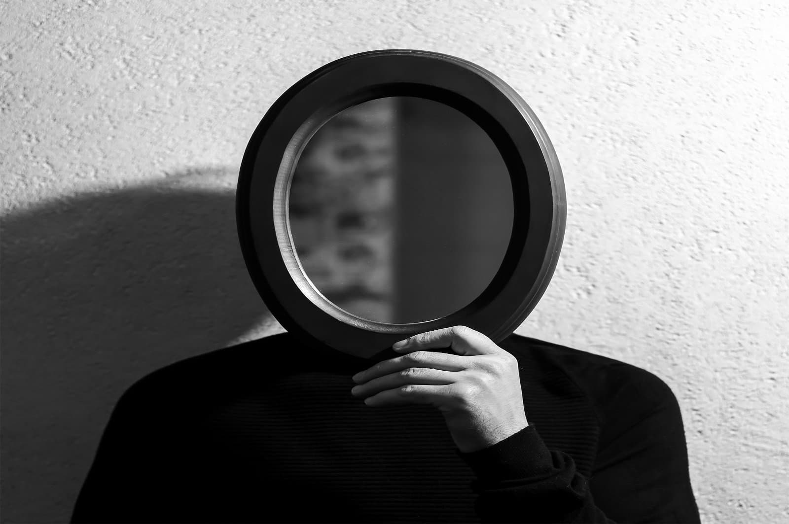 Portrait of young man holding oval mirror on face. Background of textured wall.