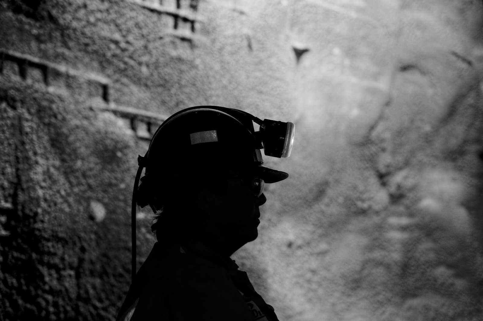 A woman working underground in a mine is silhouetted against a well lit tunnel wall. She wears a hard hat helmet with a miners lamp on it.